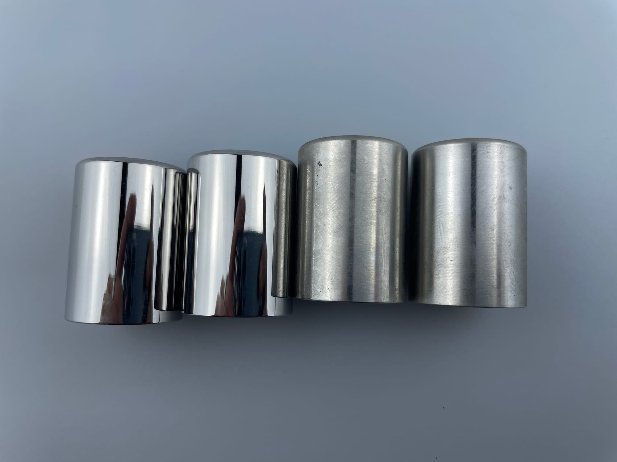 The image shows a set of four stainless steel cylinders on a white background. The cylinders are cylindrical in shape and have a glossy finish. They are arranged in a neat row, with each cylinder slig
